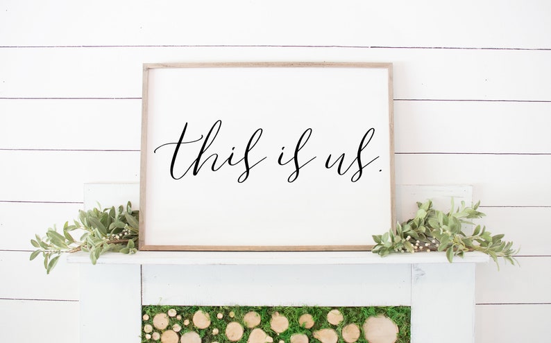 Pu&ograve; includere: Un cartello bianco incorniciato con il testo "this is us." in scrittura corsiva nera. Il cartello &egrave; appeso a una parete bianca a listelli di legno sopra un camino bianco.