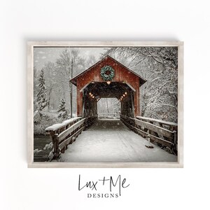 May include: A framed print of a snow-covered covered bridge. The red bridge has a Christmas wreath and lights. Snow covers the bridge and surrounding trees. The print is in a white frame. The text "Lux + Me Designs" is at the bottom.
