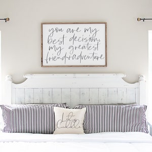 May include: A white wooden sign with black calligraphy that reads "You are my best decision, my greatest friend + adventure". The sign is hanging above a white bed with black and white striped pillows and a white pillow with black calligraphy that reads "Let's Cuddle".