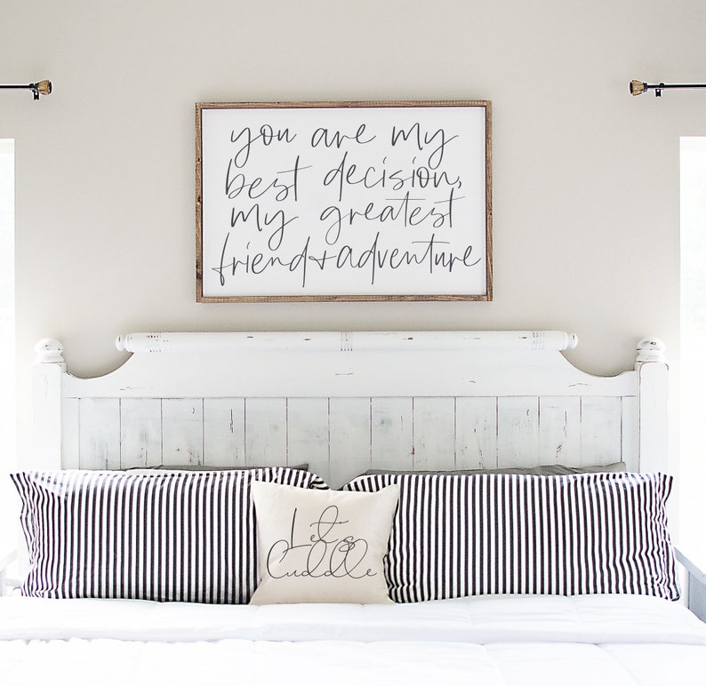 May include: A white wooden sign with black calligraphy that reads "You are my best decision, my greatest friend + adventure". The sign is hanging above a white bed with black and white striped pillows and a white pillow with black calligraphy that reads "Let's Cuddle".