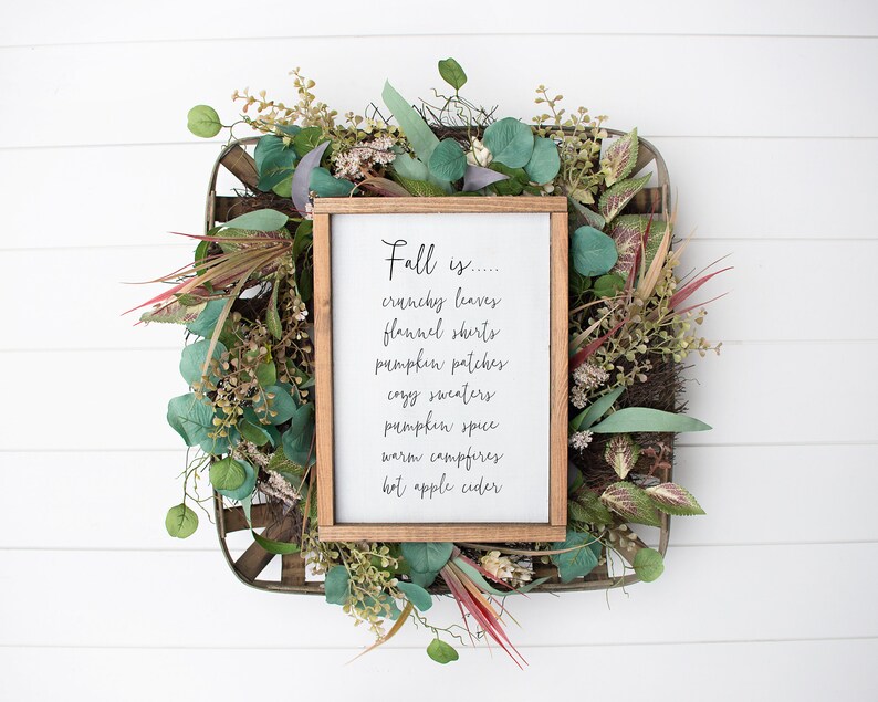 May include: A framed sign with the text "Fall is... crunchy leaves, flannel shirts, pumpkin patches, cozy sweaters, pumpkin spice, warm campfires, hot apple cider" surrounded by a wreath of green leaves.