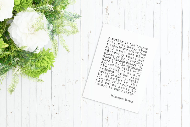 May include: A white card with black text on a white wooden background. The text reads: "A mother is the truest friend we have, when trials, heavy and sudden fall upon us; when adversity takes the place of prosperity; when friends desert us; when trouble thickens around us, still will she cling to us and endeavor by her kind precepts and counsels to dissipate the clouds of darkness, and cause peace to return to our hearts. -Washington Irving"