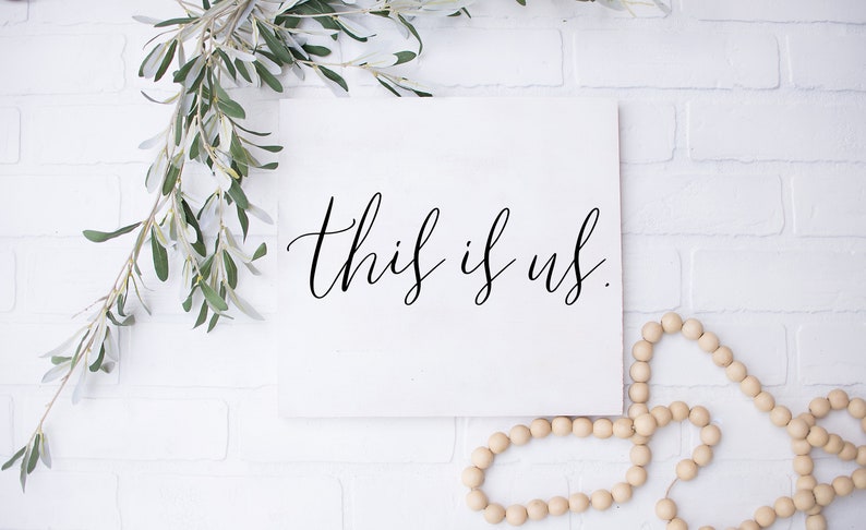 Pu&ograve; includere: Un cartello bianco con calligrafia nera che recita "this is us". Il cartello &egrave; su un muro di mattoni bianchi con una collana di perline di legno nell'angolo in basso a destra.