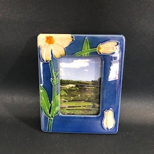 May include: A rectangular blue ceramic picture frame with a floral design. The frame features a white and yellow daisy and tulip design with green leaves. The frame holds a landscape photo. The frame is approximately 13 cm tall and 10 cm wide.