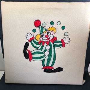 Può includere: Un cuscino quadrato color crema con un disegno di clown ricamato. Il clown indossa un abito a righe verdi e bianche, che fa giocoleria con palle verdi. Il clown ha i capelli gialli, un naso rosso e un colletto rosso. Il disegno è centrato sul cuscino.