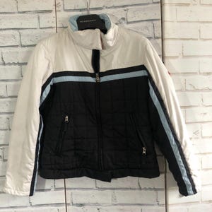 May include: A white, black, and light blue quilted jacket with a high collar and a full-length zip. The jacket has a black lower section and white upper section with light blue stripes. The brand name "BURBERRY" is visible on the hanger.