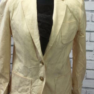 May include: A pale yellow blazer with two front pockets and gold buttons. The jacket is displayed on a mannequin, highlighting its tailored cut and classic style. The fabric looks like linen or a similar lightweight material.
