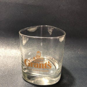 Pode incluir: Um copo de vidro transparente com formato triangular arredondado. A parte frontal do copo apresenta o logótipo dourado William Grant's e o texto Finest Scotch Whisky impresso numa fonte com serifa, centrado na parte inferior do copo.