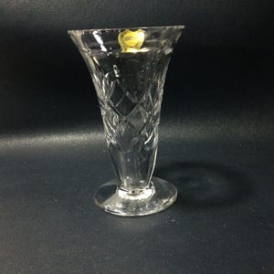 Puede incluir: Un jarrón de vidrio transparente con un diseño de cristal tallado. El jarrón tiene una parte superior acampanada y una base redonda. El jarrón tiene una etiqueta dorada con el texto "Made in England".