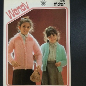 May include: Two girls wearing pink and blue fuzzy cardigans. The girl on the left is holding a wicker basket. The image is from a 1985 Wendy Mohair knitting pattern.