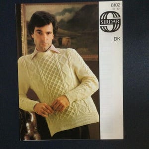 May include: A white knitted sweater with a cable knit pattern. The sweater has a round neckline and long sleeves. The pattern is a combination of diamonds and cables. The sweater is shown on a model.