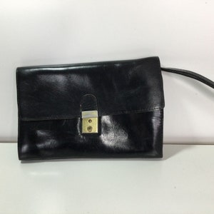 Puede incluir: Bolso de mano de cuero negro con cierre dorado. El bolso rectangular tiene una solapa y una fina correa para el hombro. El cuero parece ligeramente texturizado y el bolso está sobre un fondo blanco.