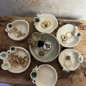 May include: Six small white ceramic dishes with green accents, each with a different piece of jewelry inside. The dishes are arranged on a wooden surface.