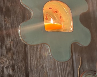May include: A teal-colored, flower-shaped wall sconce with a lit candle inside. The sconce is mounted on a weathered wooden surface. The candle flame casts a warm glow, creating a cozy ambiance. The sconce has a rustic, handmade appearance.