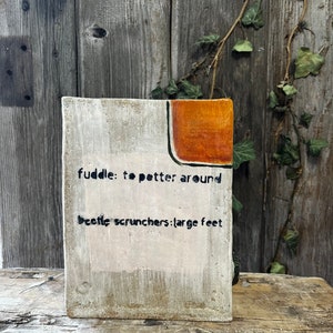 May include: A white ceramic block with a light orange and black design. The block has the text "fuddle: to potter around" and "beetle scrunchers: large feet" written on it.
