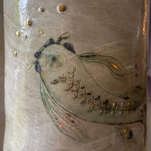 Könnte beinhalten: Keramikbecher mit handgemaltem Koi-Fisch-Design. Der Fisch ist in Grün- und Goldtönen dargestellt, mit goldenen Akzenten auf den Schuppen und Blasen. Der Becher hat einen neutralen, cremefarbenen Hintergrund mit strukturierter Oberfläche.