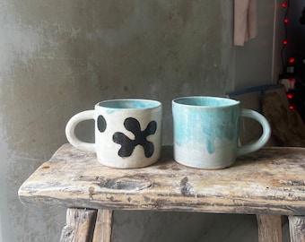 May include: Two ceramic mugs, one with a black abstract design and the other with a light blue glaze. The mugs are sitting on a wooden surface.