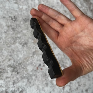 Maker of item is holding a shiny black paint brush rest between index finger and thumb.  The item has four generous spaces on which to rest paint brushes.  There is also a hole to place an incense stick.