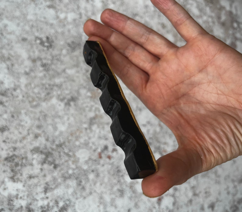 Maker of item is holding a shiny black paint brush rest between index finger and thumb.  The item has four generous spaces on which to rest paint brushes.  There is also a hole to place an incense stick.