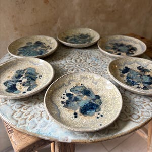 May include: Six small ceramic plates with a speckled beige glaze and abstract blue and teal designs. The plates are arranged on a decorative, round table with a floral pattern.