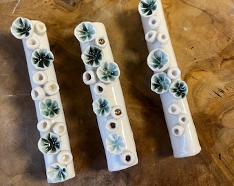 May include: Three white ceramic sticks with raised green and white floral designs and gold accents. The sticks are arranged horizontally on a wooden surface.