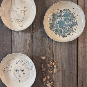 May include: Three ceramic plates with unique designs hang on a wooden wall. One plate features a line drawing of a face, another has a blue and green speckled pattern, and the third has a gold outline of a face.