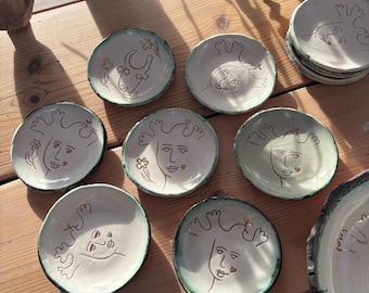May include: A collection of small, handmade ceramic plates with whimsical face designs. The plates feature a white base with a green rim and brown line art. Some plates have floral accents. The plates are arranged on a wooden surface.
