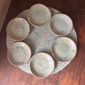 May include: A handmade ceramic serving set featuring a large, round platter with a textured floral design and six small, matching plates. The set is glazed in a speckled, muted green with brown and teal accents, perfect for appetizers or small dishes.
