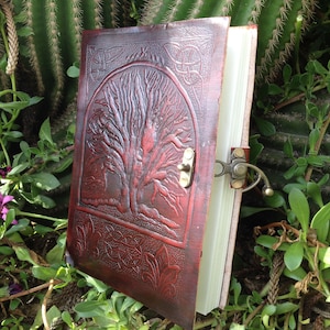 May include: A burgundy leather journal with a tree of life design embossed on the front cover. The journal has a clasp closure and is closed.