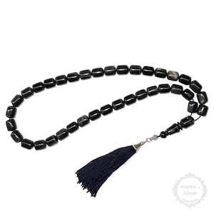 Peut inclure: Chapelet de prière noir avec un pompon bleu foncé. Les perles sont en matériau foncé opaque et sont enfilées sur une chaîne argentée. Le pompon est attaché à la chaîne par un fermoir argenté.
