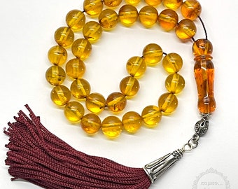 May include: A golden amber prayer beads necklace with a tassel and a silver charm. The beads are round and have a translucent appearance. The tassel is a deep burgundy color.