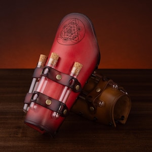May include: A red leather bracer featuring a dark gradient edge, a black geometric circular engraving, and two dark brown leather straps holding three clear glass vials with cork stoppers. A second brown leather bracer is partially visible behind it.