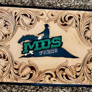 May include: A rectangular, light brown leather piece with intricate floral and scrollwork carvings. The center features a green and white logo with a rider on a horse and the text "MDS FARMS". The border is dark blue.