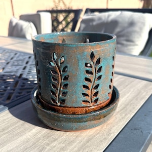 May include: A cylindrical ceramic planter with a matching saucer, featuring a teal and brown glaze. The exterior of the planter is decorated with vertical cutouts in a leaf-like pattern, revealing the interior through the clay walls.