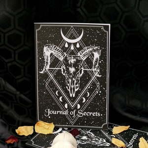 Puede incluir: Dos diarios negros con un dise&ntilde;o de calavera blanca y luna creciente. El texto de la portada dice "Journal of Secrets". Un cr&aacute;neo de animal blanco est&aacute; en primer plano.