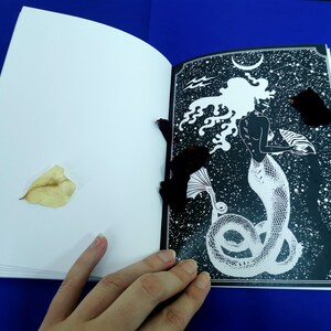 Puede incluir: Un libro abierto con una ilustraci&oacute;n en blanco y negro de una sirena con el pelo largo y fluido y una cola. La sirena sostiene una concha en su mano. El fondo es un cielo estrellado en blanco y negro.