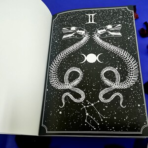 Puede incluir: Una ilustraci&oacute;n en blanco y negro de una serpiente esquel&eacute;tica con una luna y estrellas sobre un fondo negro. La imagen es parte de un libro con una portada blanca.