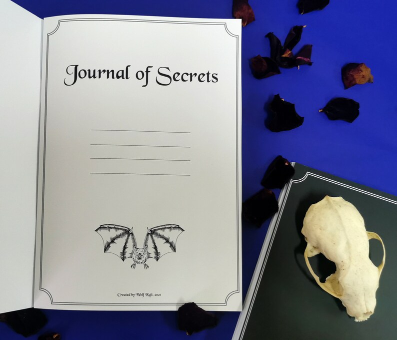 Puede incluir: Un diario blanco con un borde negro y el texto "Journal of Secrets" en la portada. El diario tiene una ilustraci&oacute;n en blanco y negro de un murci&eacute;lago en la parte inferior de la portada. El diario est&aacute; abierto a una p&aacute;gina en blanco.