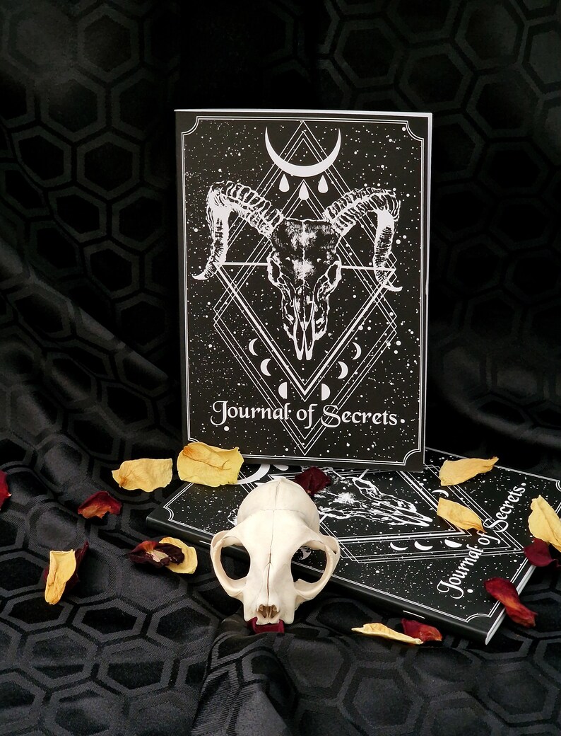 Puede incluir: Dos diarios negros con un dise&ntilde;o de calavera blanca y luna creciente. El texto de la portada dice "Journal of Secrets". Un cr&aacute;neo de animal blanco est&aacute; en primer plano.
