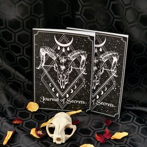 Puede incluir: Dos diarios negros y blancos con un dise&ntilde;o de calavera y luna creciente. El texto de la portada dice "Journal of Secrets". Los diarios est&aacute;n sobre un fondo de tela negra con p&eacute;talos de rosa.