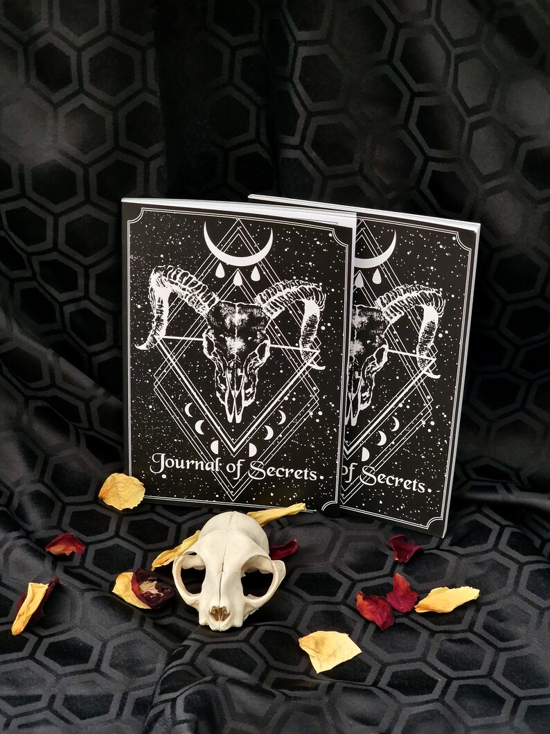 Puede incluir: Dos diarios negros y blancos con un dise&ntilde;o de calavera y luna creciente. El texto de la portada dice "Journal of Secrets". Los diarios est&aacute;n sobre un fondo de tela negra con p&eacute;talos de rosa.