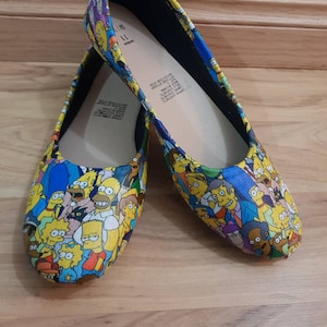 Puede incluir: Un par de zapatos planos amarillos, azules y negros con un estampado de los personajes de Los Simpson. Los zapatos tienen un forro negro y una plantilla blanca. La plantilla tiene la talla 38 escrita.