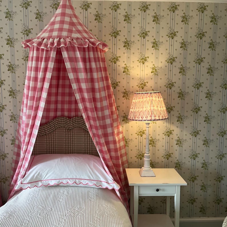 May include: A pink and white gingham canopy bed with a white pillow and white and pink patterned bedding. A white bedside table with a white lamp with a pink and white patterned shade.