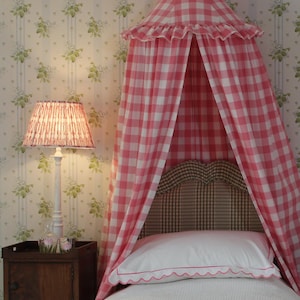 May include: A pink and white gingham canopy bed with a white ruffled trim. The bed has a white headboard and a white bedspread with a pink trim. The bed is in a room with floral wallpaper and a white lamp with a pink shade.