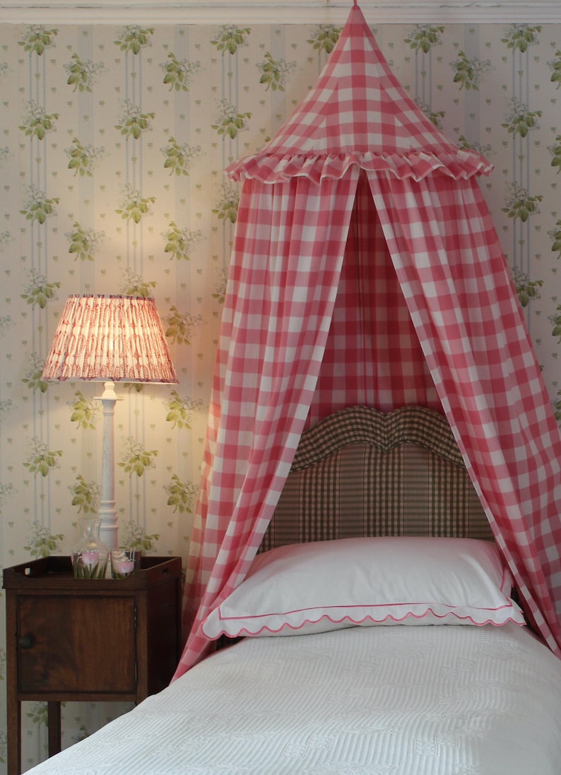 May include: A pink and white gingham canopy bed with a white ruffled trim. The bed has a white headboard and a white bedspread with a pink trim. The bed is in a room with floral wallpaper and a white lamp with a pink shade.