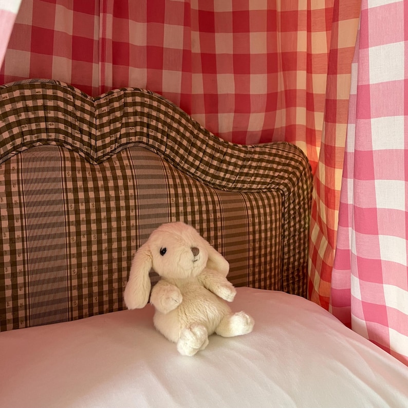 May include: A white plush bunny rabbit toy sits on a white bed with a brown and white checked headboard. The bed is covered with a pink and white gingham canopy.