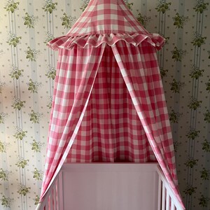 May include: A pink and white gingham canopy hangs over a white cot with two stuffed toys inside. The canopy has a ruffled edge.