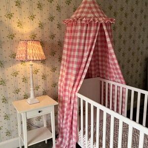 May include: A white cot with a pink and white gingham canopy draped over it. The cot has a white mattress and a white blanket with a floral pattern. A white bedside table with a lamp is next to the cot.