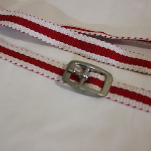 Puede incluir: Primer plano de una correa a rayas rojas y blancas con una hebilla de metal plateado. La correa está hecha de un material tejido y parece nueva. El fondo es una superficie blanca lisa.