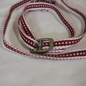 Peut inclure: Une ceinture tissée avec un motif répétitif de rayures rouges et blanches. La ceinture est dotée d'une boucle en métal argenté. La ceinture est posée sur une surface blanche.
